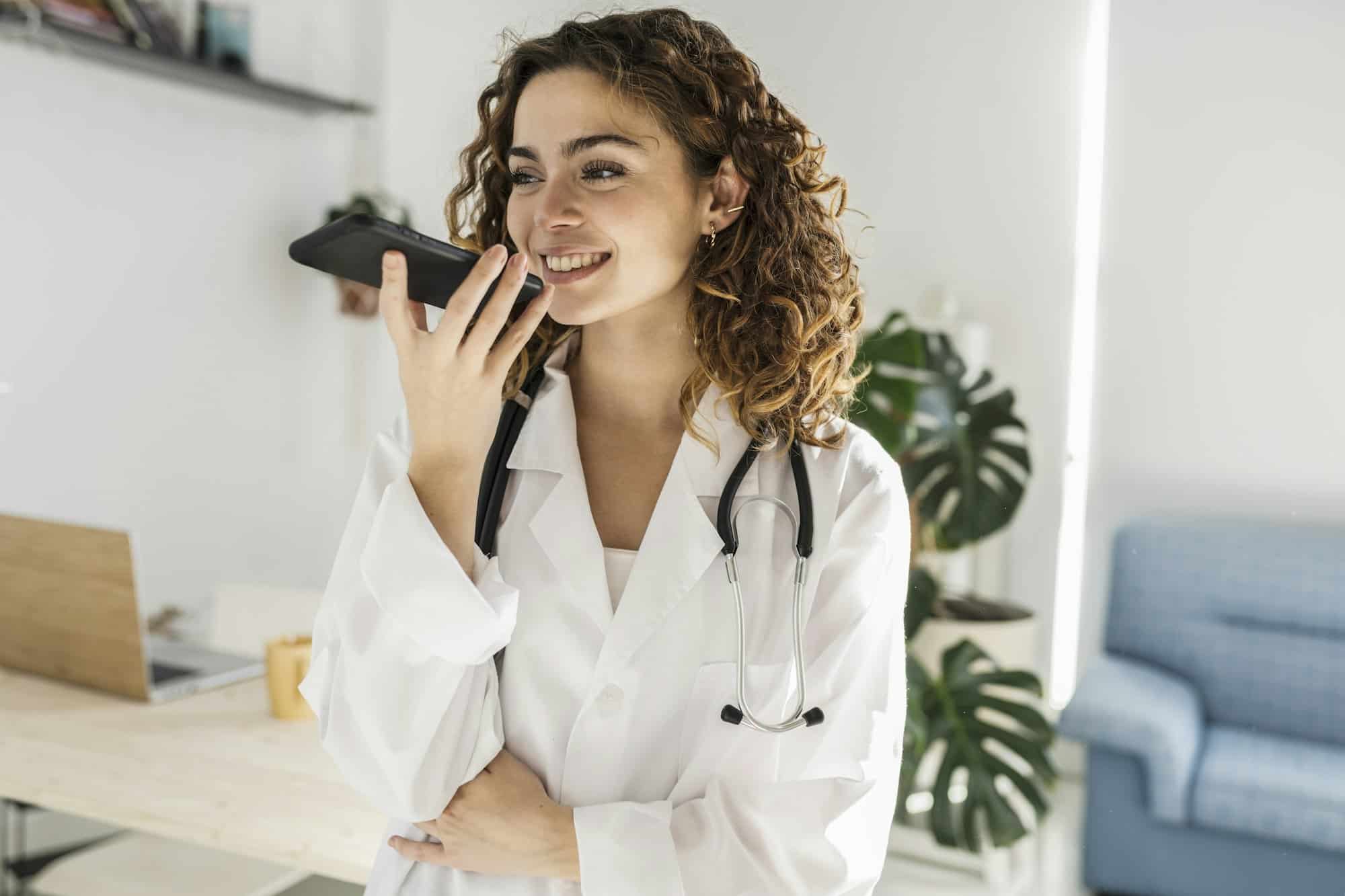 Young curly haired female doctor sending an audio message to a patient with her smartphone